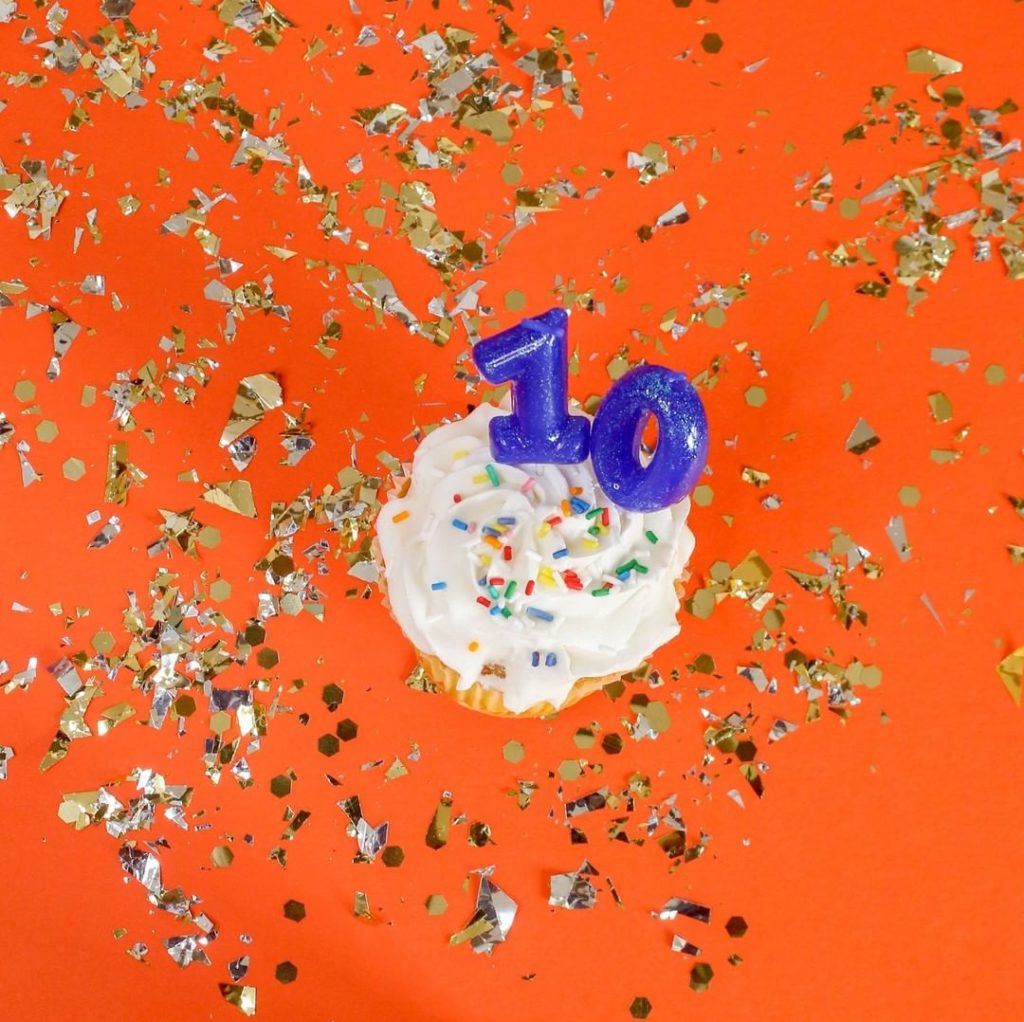 Cupcake with number candles that say "10" on a backround of solid colored orange with gold confetti